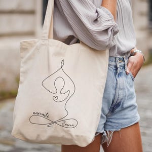 May include: Beige tote bag featuring a black line drawing of a person in a yoga pose with a heart. The words "namaste" and "Emma" are written in a script font. The bag has long handles.