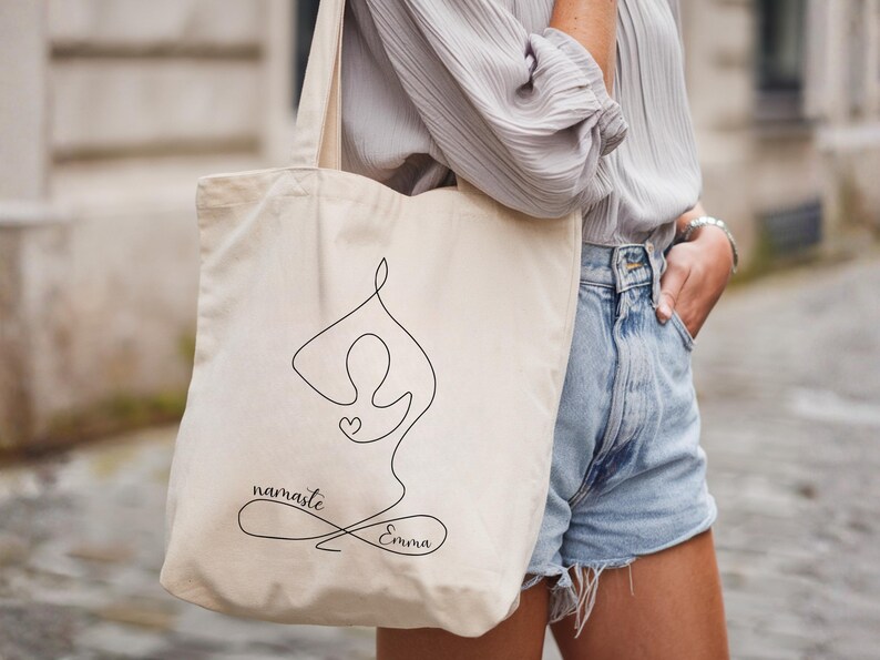 May include: Beige tote bag featuring a black line drawing of a person in a yoga pose with a heart. The words "namaste" and "Emma" are written in a script font. The bag has long handles.