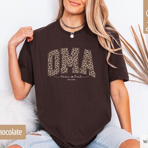May include: Dark chocolate brown t-shirt with the word "OMA" in a leopard print design. Below the word "OMA" is the text "Tamar & Noah" and "EST 2026". The shirt is made of 100% cotton.