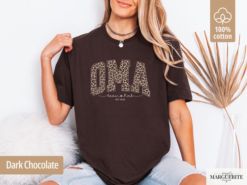 May include: Dark chocolate brown t-shirt with the word "OMA" in a leopard print design. Below the word "OMA" is the text "Tamar & Noah" and "EST 2026". The shirt is made of 100% cotton.