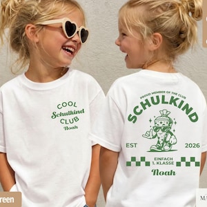 May include: A white t-shirt featuring green text and graphics. The front displays 'COOL Schulkind CLUB Noah'. The back shows a cartoon character in a cone, the text 'PROUD MEMBER OF THE CLUB SCHULKIND', 'EST 2026', 'EINFACH 1. KLASSE', and 'Noah'.
