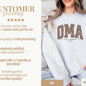 May include: A light gray sweatshirt featuring the word "OMA" in a leopard print design. Below the word "OMA" is the text "Emma & Noah" and "EST 2020". The sweatshirt also has customer reviews on the left side.