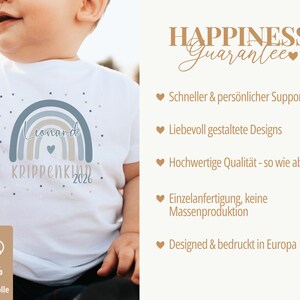 May include: A white short-sleeved t-shirt featuring a graphic of a muted blue and beige rainbow with the text Leonard, KRIPPENKIND, and 2026. The shirt includes a small heart icon and scattered dots. A label indicates 100% organic cotton.