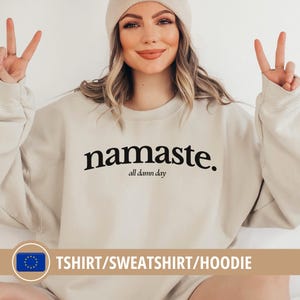 Könnte beinhalten: Beiges Sweatshirt mit dem schwarzen Schriftzug "namaste" und "all damn day" darunter. Das langärmlige Oberteil hat eine neutrale Farbe. Eine beige Mütze ist ebenfalls zu sehen.
