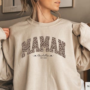 Op de afbeelding: Zandkleurige sweatshirt met het woord "MAMA" in luipaardprint over de borst. Onder het woord "MAMA" staat de naam "charlotte" en het jaar "EST 2026". De sweatshirt heeft lange mouwen en een ronde hals.