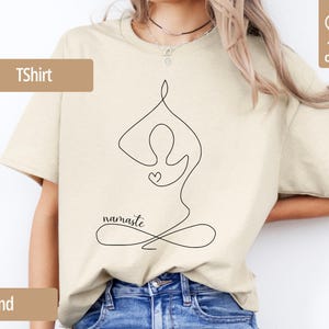 May include: Sand-colored T-shirt with a black line drawing of a person in a yoga pose, a heart, and the word "namaste." The shirt is made of 100% cotton.