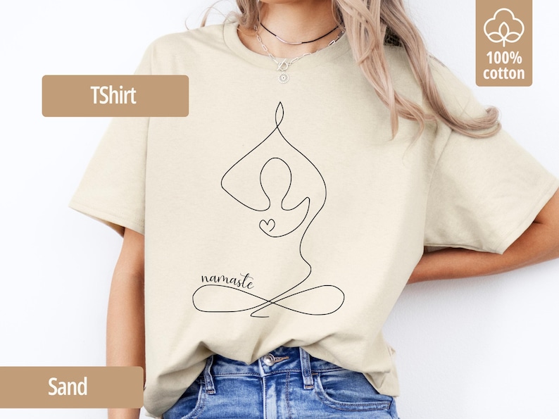 May include: Sand-colored T-shirt with a black line drawing of a person in a yoga pose, a heart, and the word "namaste." The shirt is made of 100% cotton.