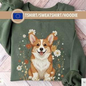 Può includere: Felpa verde militare con un'illustrazione di un cane corgi sorridente circondato da fiori. Il testo "TSHIRT/SWEATSHIRT/HOODIE" è visualizzato in alto. Un capo comodo e casual.