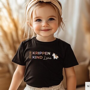 May include: A black short-sleeved t-shirt featuring the text 2026 KRIPPEN KIND Lena in muted earth tones, accompanied by a small graphic of a white unicorn with a gold horn and a saddle, centered on the chest of the garment.