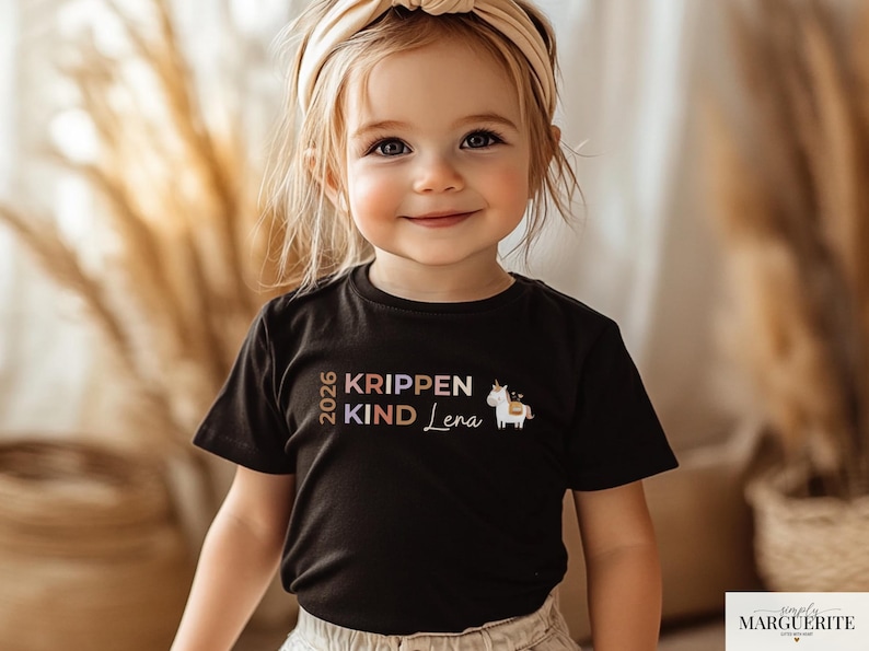 May include: A black short-sleeved t-shirt featuring the text 2026 KRIPPEN KIND Lena in muted earth tones, accompanied by a small graphic of a white unicorn with a gold horn and a saddle, centered on the chest of the garment.
