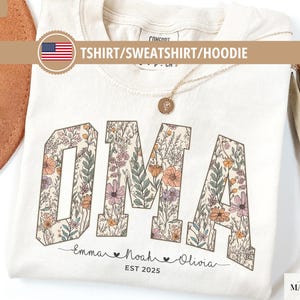 May include: Ivory-colored T-shirt with the word "OMA" in large, floral-patterned letters. Below the word are the names "Emma", "Noah", and "Olivia", with the year "EST 2025". The shirt is labeled "TSHIRT/SWEATSHIRT/HOODIE".