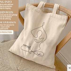 May include: Beige canvas tote bag with black line art of a yoga pose, a heart, and the words "namaste Sophie." The bag has two handles and is sitting on a woven chair. The bag is made of a natural material.