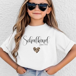 May include: A white short-sleeved t-shirt featuring the black cursive text Schulkind, the name Olivia 2026 in smaller black script, and a small heart graphic with a brown and black leopard print pattern centered below the text.