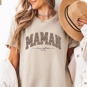 Op de afbeelding: Een zandkleurig t-shirt met het woord "MAMAN" in een luipaardprint. Daaronder staat de naam "Lucian Rose" en het jaar "EST 2024". De persoon draagt een strohoed en een witte jas.