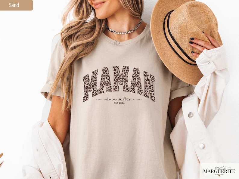 Op de afbeelding: Een zandkleurig t-shirt met het woord "MAMAN" in een luipaardprint. Daaronder staat de naam "Lucian Rose" en het jaar "EST 2024". De persoon draagt een strohoed en een witte jas.