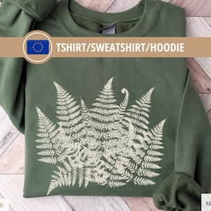 Pode incluir: Camisola verde militar com um desenho de samambaia branca. O texto "TSHIRT/SWEATSHIRT/HOODIE" é exibido em uma faixa. A camisola está dobrada sobre uma superfície de madeira.