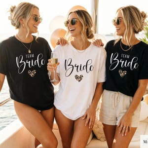 Op de afbeelding: Drie vrouwen dragen zwarte en witte t-shirts met de woorden "Team Bride" en "Bride" in cursief schrift. Elk shirt heeft een hart met luipaardprint. De vrouwen lachen en dragen zonnebrillen.