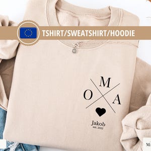 May include: A sand-colored sweatshirt with the word "OMA" crossed by lines, a black heart, and the name "Jakob est. 2022" printed below. The shirt is folded on a white surface, with a necklace and a "TSHIRT/SWEATSHIRT/HOODIE" banner.