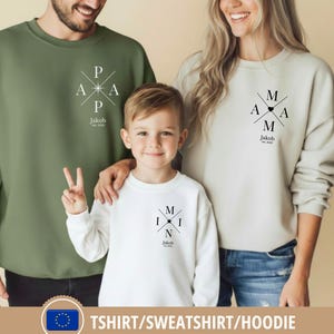 Op de afbeelding: Familie bijpassende sweatshirts in olijfgroen, wit en crème. Elk sweatshirt heeft een uniek ontwerp met de woorden "PAPA", "MAMA" en "MINI" in een kruisvormig ontwerp, samen met de naam "Jakob" en het jaar "est. 2022".