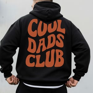 May include: Black hoodie with the words "COOL DADS CLUB" in orange, retro-style lettering across the back. The hoodie has a hood and long sleeves. The person is wearing a white undershirt.