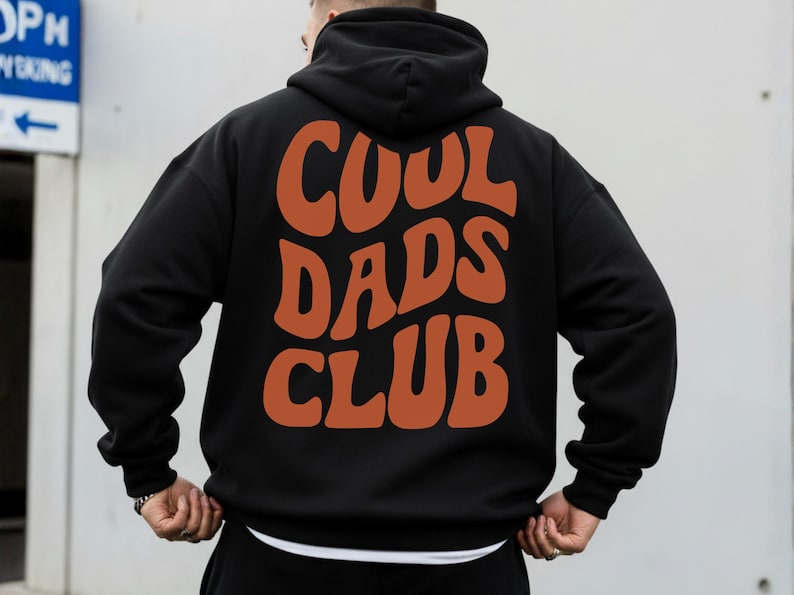 May include: Black hoodie with the words "COOL DADS CLUB" in orange, retro-style lettering across the back. The hoodie has a hood and long sleeves. The person is wearing a white undershirt.