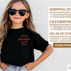 May include: A black t-shirt featuring a red circular graphic on the chest with the text COOL SCHULKIND CLUB and the name Emma printed below. The shirt has short sleeves and a crew neckline, worn with heart-shaped sunglasses.