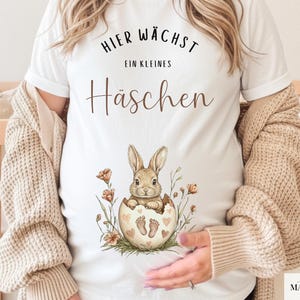 Peut inclure: T-shirt blanc avec le texte allemand "Hier wächst ein kleines Häschen" et une illustration aquarelle d'un lapin sortant d'un œuf. L'œuf a des motifs de cœurs et d'empreintes de pas, entouré de fleurs.