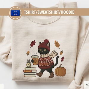 Può includere: Felpa color sabbia con un disegno ricamato di un gatto nero con cappello, sciarpa e maglione, che tiene una tazza di caffè. Il disegno comprende foglie autunnali, ghiande, una zucca e libri. Il testo "TSHIRT/SWEATSHIRT/HOODIE" è in alto.