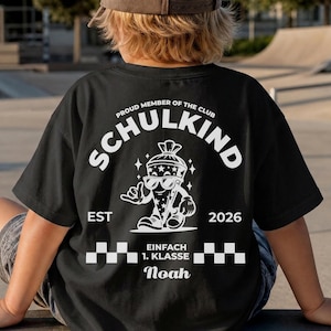 May include: A black t-shirt featuring a white graphic on the back. The design includes the text "PROUD MEMBER OF THE CLUB SCHULKIND", "EST 2026", and "EINFACH 1. KLASSE" with the name "Noah". A cartoon character wearing sunglasses is centered.