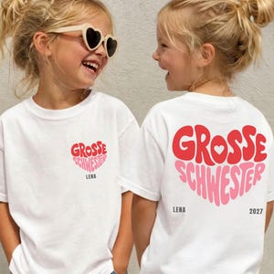 May include: A white t-shirt featuring a red and pink heart-shaped graphic on the front chest with the text "GROSSE SCHWESTER" and "LENA". The back displays a larger version of the same graphic with "LENA" and "2027" printed below.