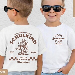 May include: A white t-shirt featuring brown graphic prints. The back displays a cartoon school cone character with sunglasses, the text SCHULKIND, EST 2026, EINFACH 1. KLASSE, and the name Theodor. The front shows the text COOL Schulkind CLUB Theodor.