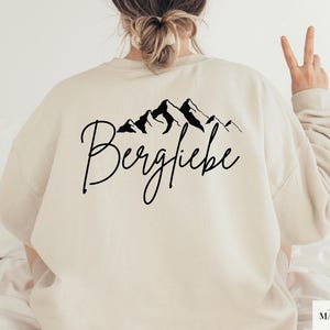 May include: A cream-colored sweatshirt with a black mountain range graphic and the word "Bergliebe" in cursive. The sweatshirt is a long-sleeved, casual top.