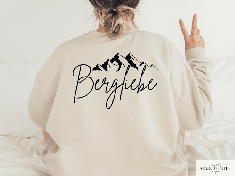 May include: A cream-colored sweatshirt with a black mountain range graphic and the word "Bergliebe" in cursive. The sweatshirt is a long-sleeved, casual top.