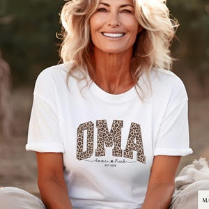 May include: White t-shirt featuring the word "OMA" in a leopard print design. Underneath, the text reads "Emma & Noah" with a heart symbol and the year "EST 2020". The shirt is worn by a person.