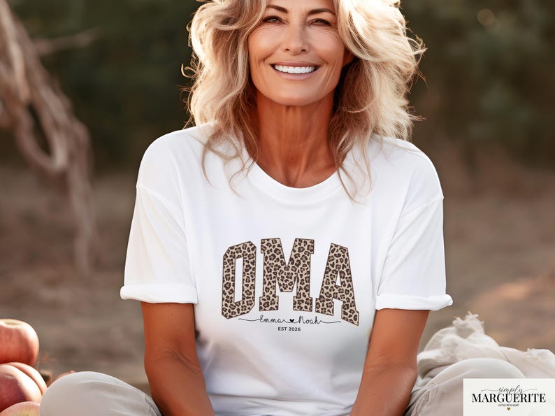 May include: White t-shirt with the word "OMA" in leopard print. Below the word "OMA" is the text "Emma & Noah" with a heart symbol and the year "EST 2020". The shirt is worn by a person.