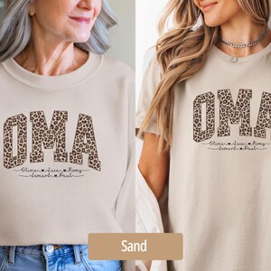 May include: Sand-colored sweatshirt and t-shirt featuring the word "OMA" in a leopard print design. Below the word are names and hearts. The sweatshirt has long sleeves, while the t-shirt has short sleeves.