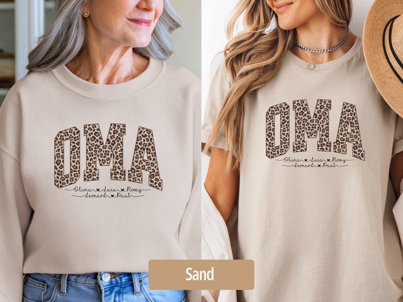 May include: Sand-colored sweatshirt and t-shirt featuring the word "OMA" in a leopard print design. Below the word are names and hearts. The sweatshirt has long sleeves, while the t-shirt has short sleeves.