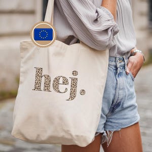 Könnte beinhalten: Beige Tragetasche mit dem Wort "hej" im Leopardenmuster. Die Tasche wird von einer Person getragen, die ein hellgraues Hemd und Jeansshorts trägt. Ein kleiner Aufkleber mit der Flagge der Europäischen Union ist angebracht.