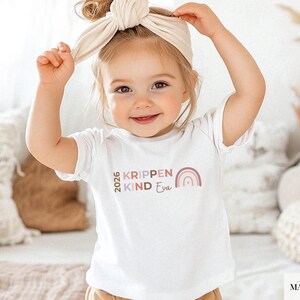 May include: A white short-sleeved t-shirt featuring the text 2026 KRIPPEN KIND Eva in muted brown, purple, and pink tones, accompanied by a small rainbow graphic. The design is printed on the chest of the shirt in a simple, clean font style.