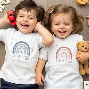 May include: Two white short-sleeved t-shirts featuring a rainbow graphic. The left shirt has a blue-toned rainbow with the text "Leonard" and "KRIPPENKIND 2026". The right shirt has a pink-toned rainbow with the text "Leona" and "KRIPPENKIND 2026".
