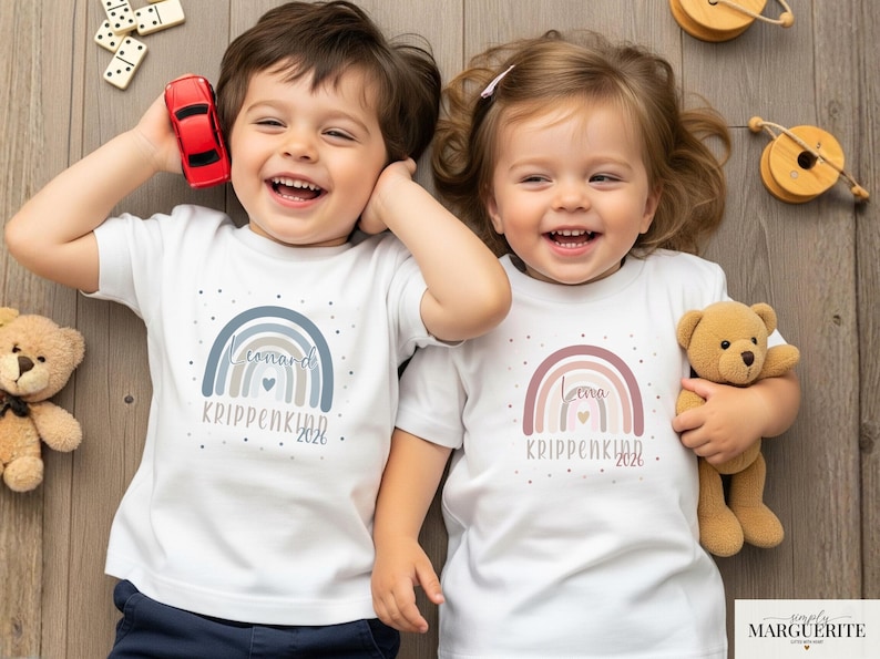 May include: Two white short-sleeved t-shirts featuring a rainbow graphic. The left shirt has a blue-toned rainbow with the text "Leonard" and "KRIPPENKIND 2026". The right shirt has a pink-toned rainbow with the text "Leona" and "KRIPPENKIND 2026".