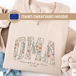 May include: A beige sweatshirt featuring the word "OMA" in a floral design. Below the word are the names "Emma Noah Olivia" and the year "EST 2025". The top is folded on a denim surface.