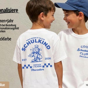 May include: A white t-shirt featuring blue graphics. The back displays a cartoon school cone character wearing sunglasses with the text SCHULKIND, EST 2026, and EINFACH 1. KLASSE. The front left chest features the text COOL SCHULKIND CLUB and Theodor.