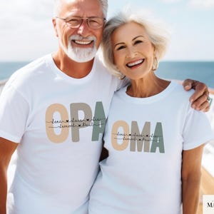 Może przedstawiać: Dwie osoby ubrane w białe koszulki z napisami "OPA" i "OMA" w stylizowanej czcionce. Koszulki mają imiona i serca pod spodem. Tłem jest niebieski ocean i biała łódź.