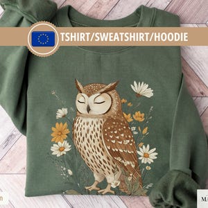 Könnte beinhalten: Militärgrünes Sweatshirt mit einer detaillierten Illustration einer braunen Eule mit geschlossenen Augen, umgeben von gelben und weißen Blumen. Der Text "TSHIRT/SWEATSHIRT/HOODIE" ist über der Eule zu sehen. Das Sweatshirt liegt auf einer Holzoberfläche.