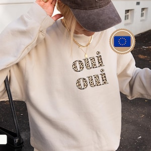 Könnte beinhalten: Ein sandfarbener Pullover mit den Worten "Oui Oui" in einem Leopardenmuster. Der Pullover hat eine neutrale Farbe und eine lockere Passform. Das Bild enthält auch eine schwarze Tasche und eine braune Kappe.