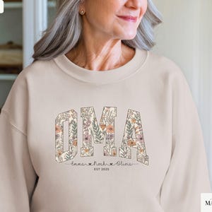 May include: Sand-colored sweatshirt with the word "OMA" in a floral design. Below the word are the names "Emma", "Noah", and "Olivia" with hearts, and the year "EST 2025". The sweatshirt is a crew neck style.