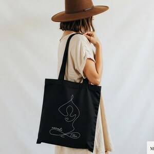May include: Black tote bag featuring a white line drawing of a yoga pose with a heart. The words "namaste" and "Chloe" are written in cursive below the figure. The bag is carried over the shoulder.