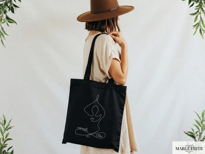 May include: Black tote bag featuring a white line drawing of a yoga pose with a heart. The words "namaste" and "Chloe" are written in cursive below the figure. The bag is carried over the shoulder.