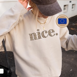 Può includere: Una felpa color sabbia con la scritta "nice" in un motivo leopardato. La felpa ha maniche lunghe e girocollo. L'immagine mostra anche una borsa nera e un berretto da baseball marrone.